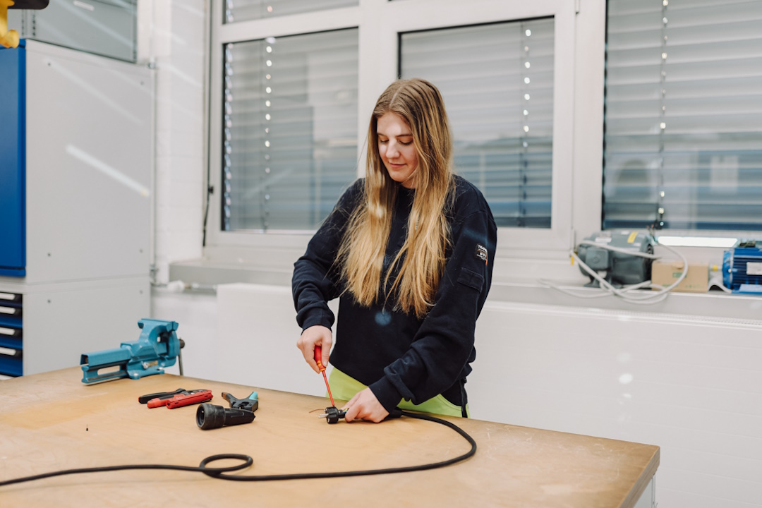 Eine junge Frau mit langen blonden Haaren arbeitet an einem Werktisch in einem hellen Werkraum. Mit einem Schraubendreher bearbeitet sie ein Kabel oder technisches Bauteil. Auf dem Tisch liegen weitere Werkzeuge, im Hintergrund sind Fenster und Werkstatte