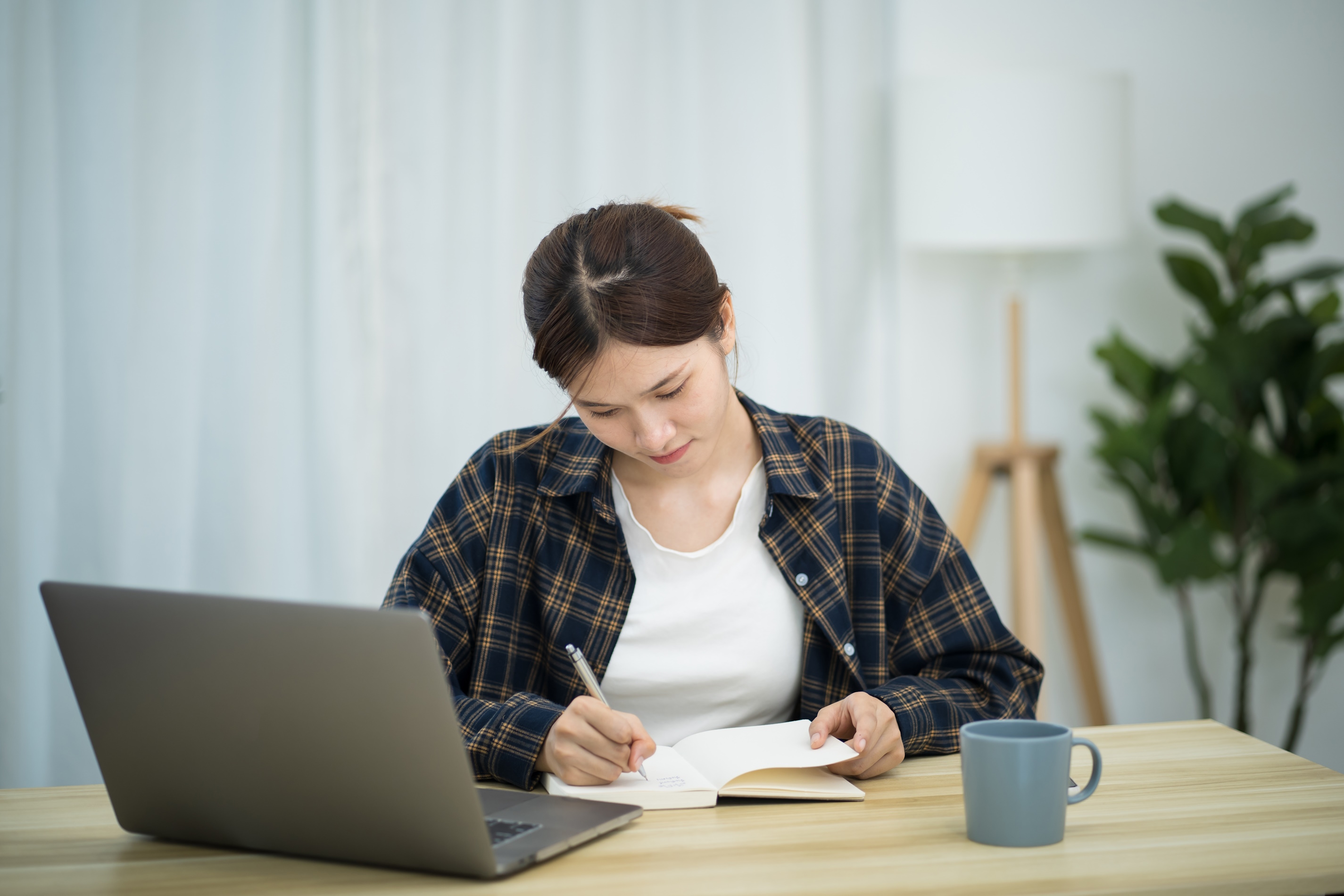 Person schreibt in ein Notizbuch am Schreibtisch; daneben stehen Laptop und Tasse.