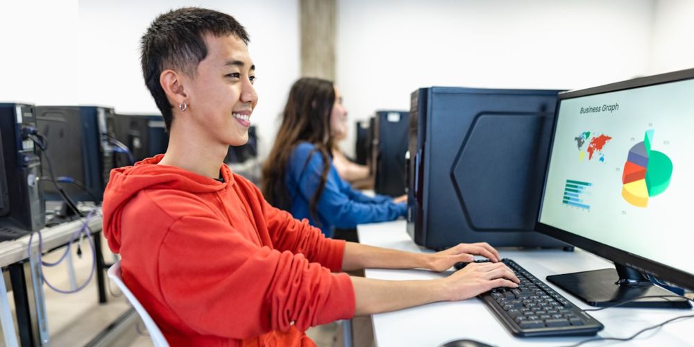 Eine Person in einem roten Kapuzenpullover sitzt an einem Schreibtisch vor einem Desktop-Computer und lächelt beim Arbeiten. Auf dem Bildschirm sind verschiedene Diagramme und Grafiken zu sehen. Im Hintergrund arbeiten weitere Personen an Computern.