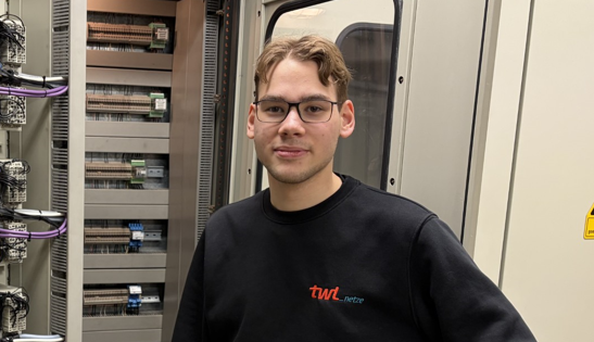 Porträt eines Auszubildenden vor einem Schaltschrank, in schwarzem Pulli mit orangenen Aufdruck TWL.