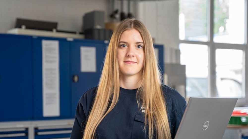 Junge Frau mit langen blonden Haaren steht in einer Werkstatt oder einem technischen Arbeitsumfeld vor blauen Werkzeugschränken und hält einen geöffneten Laptop in der Hand. Sie blickt direkt in die Kamera.