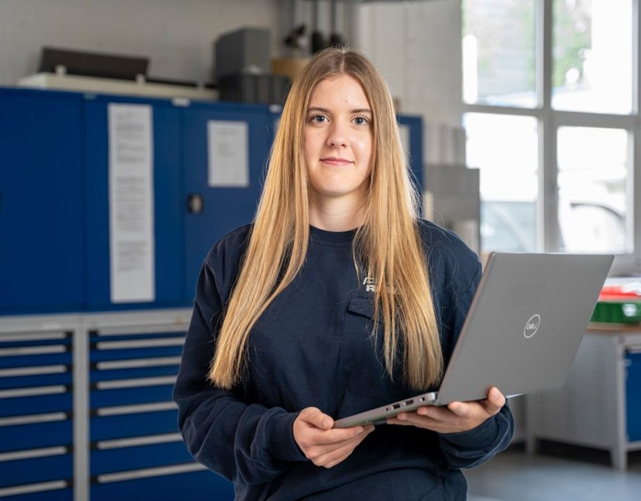 Eine junge Frau mit langen blonden Haaren steht in einer Werkstatt oder einem technischen Arbeitsraum und hält ein geöffnetes Laptop in den Händen. Sie trägt ein dunkles Sweatshirt und blickt direkt in die Kamera. Im Hintergrund sind blaue Werkstattschrän