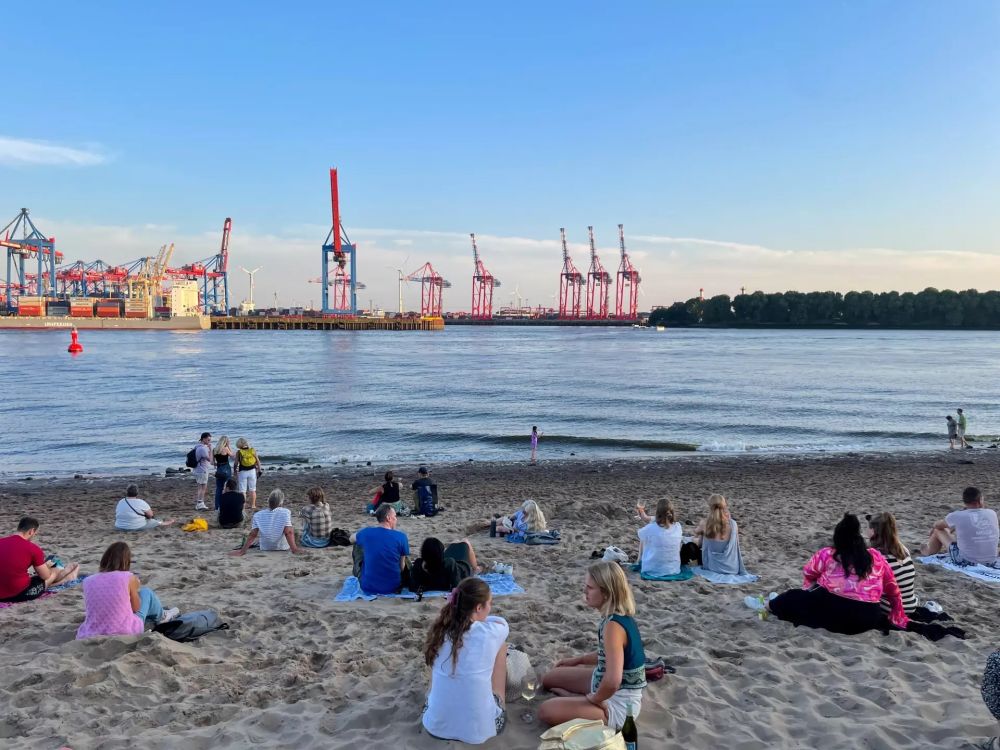 Mehrere Personen sitzen auf einem sandigen Ufer vor einer breiten Wasserfläche; am gegenüberliegenden Ufer stehen mehrere rote Hafenkräne