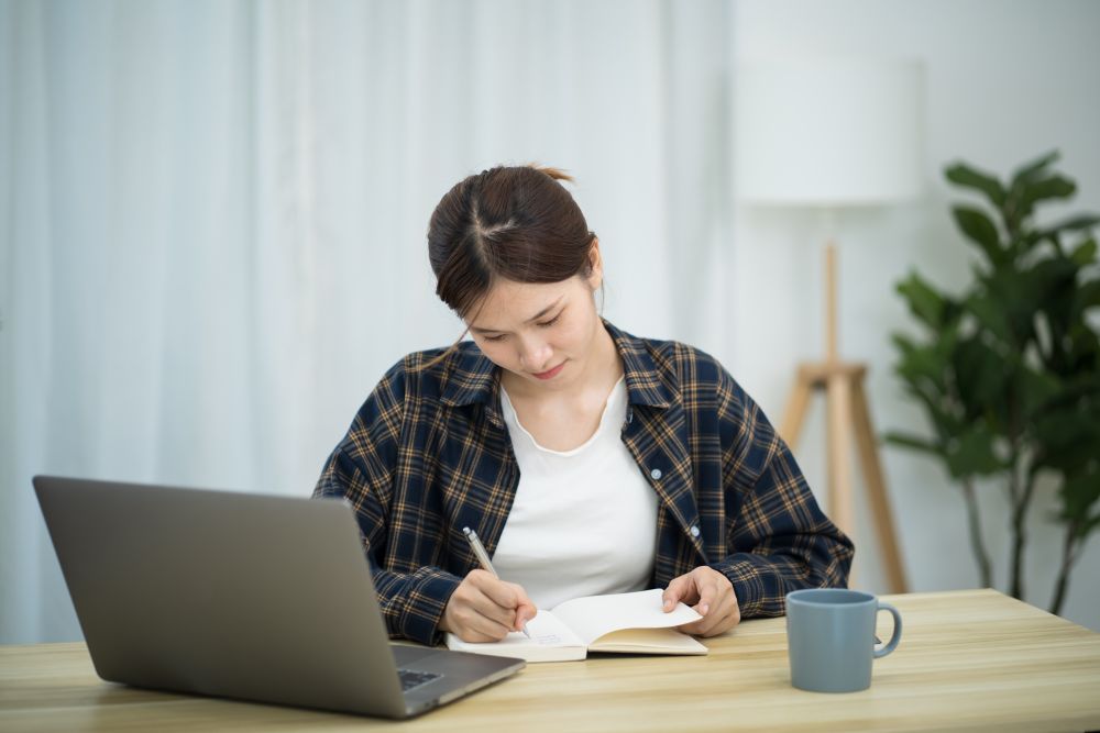 Person schreibt in ein Notizbuch am Schreibtisch; daneben stehen Laptop und Tasse.