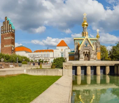 Ansicht der Darmstädter Künstlerkolonie mit golden verzierter Kapelle, Wasserbecken und dem Hochzeitsturm im Hintergrund.